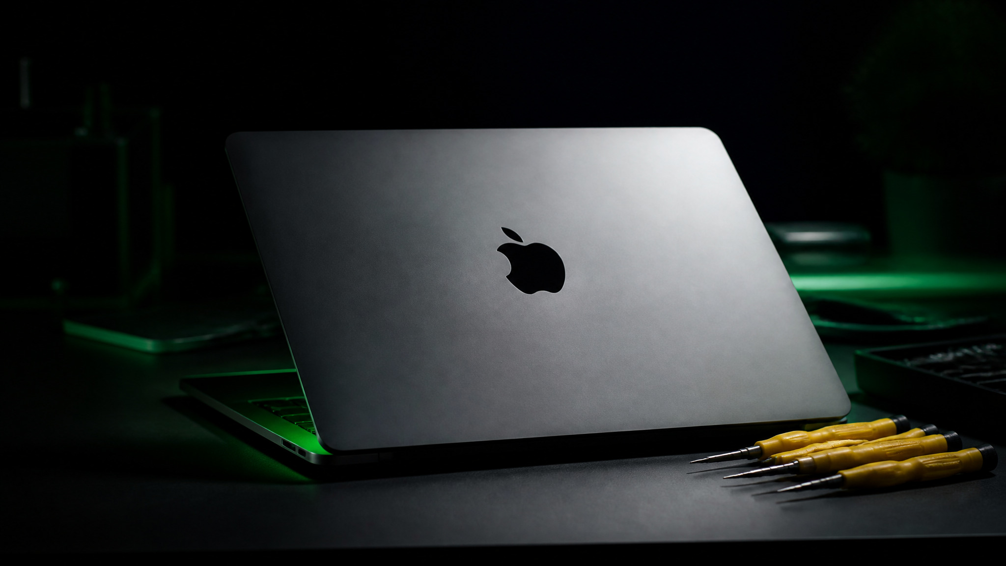 MacBook Pro slightly open on dark bench with Apple logo on lid and yellow Wiha precision tools