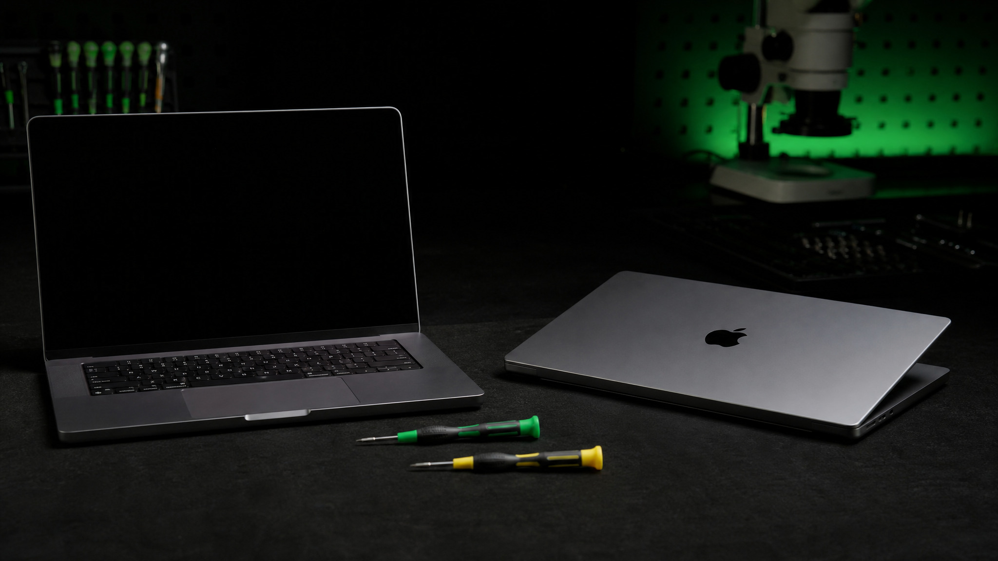 Two MacBook Pros on dark bench with both Apple logos visible and green and yellow Wiha tools between them