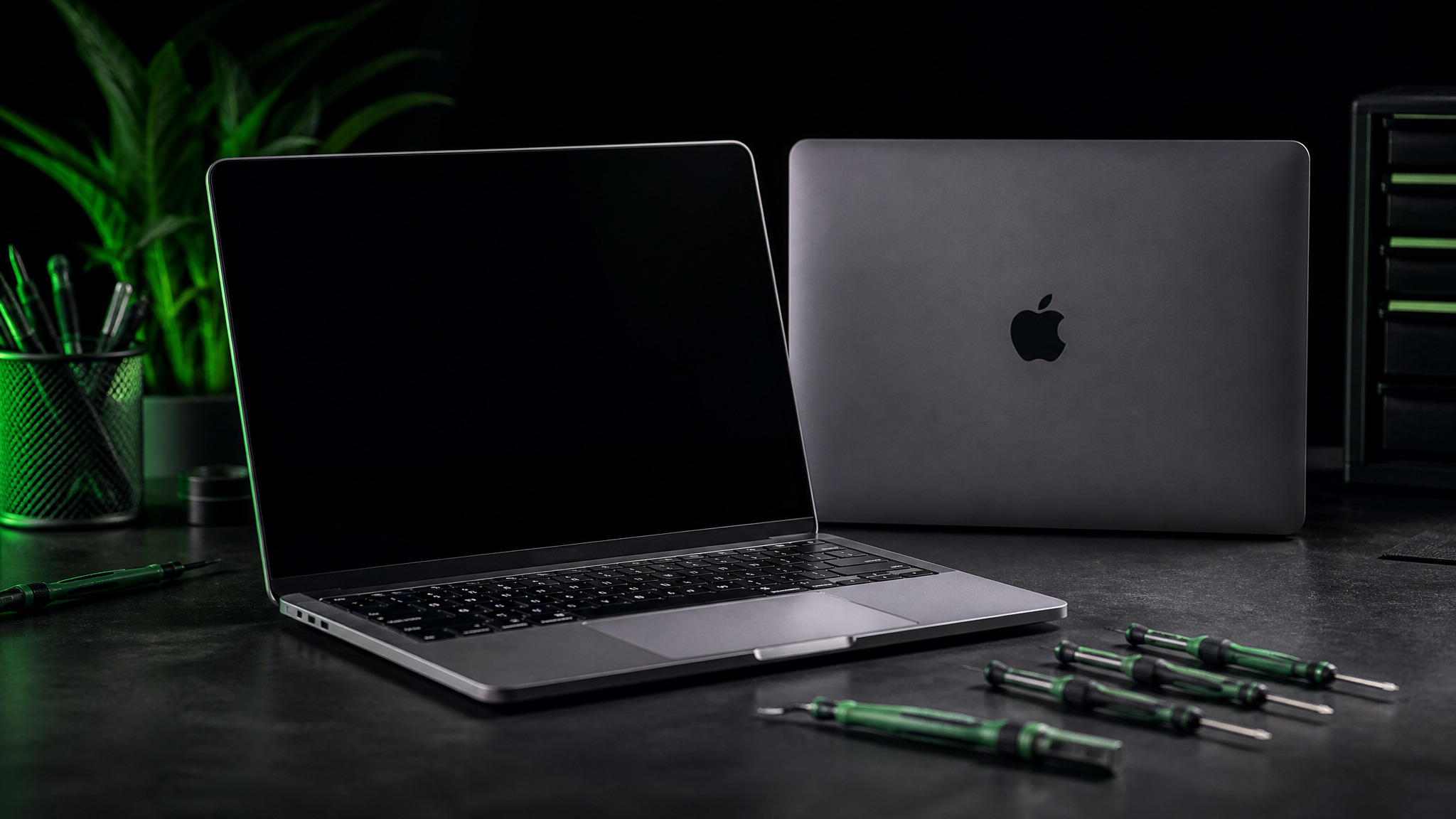 MacBook Pro open on dark bench with second MacBook showing Apple logo behind and green Wiha drivers