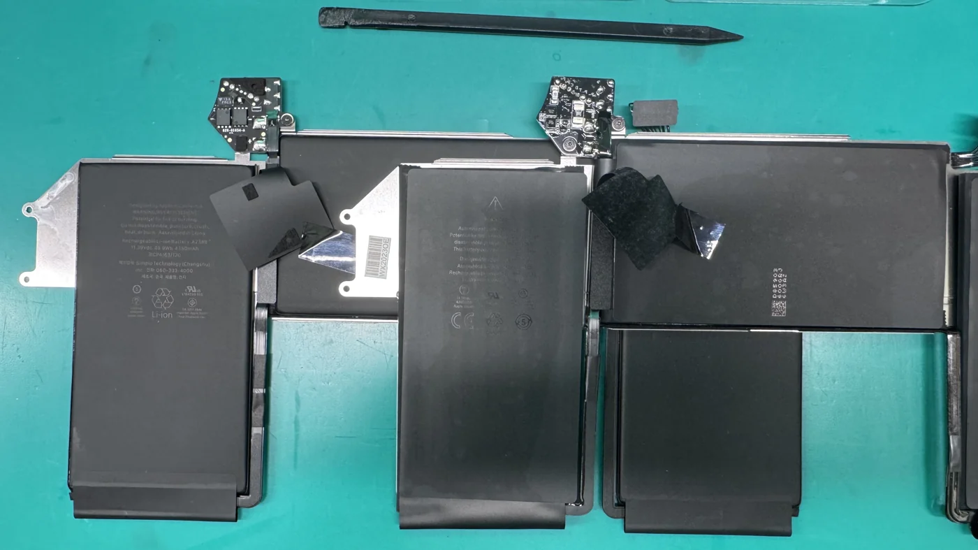 A genuine Apple MacBook battery and a counterfeit non-OEM battery laid out side by side for comparison on the WarriorMac workbench