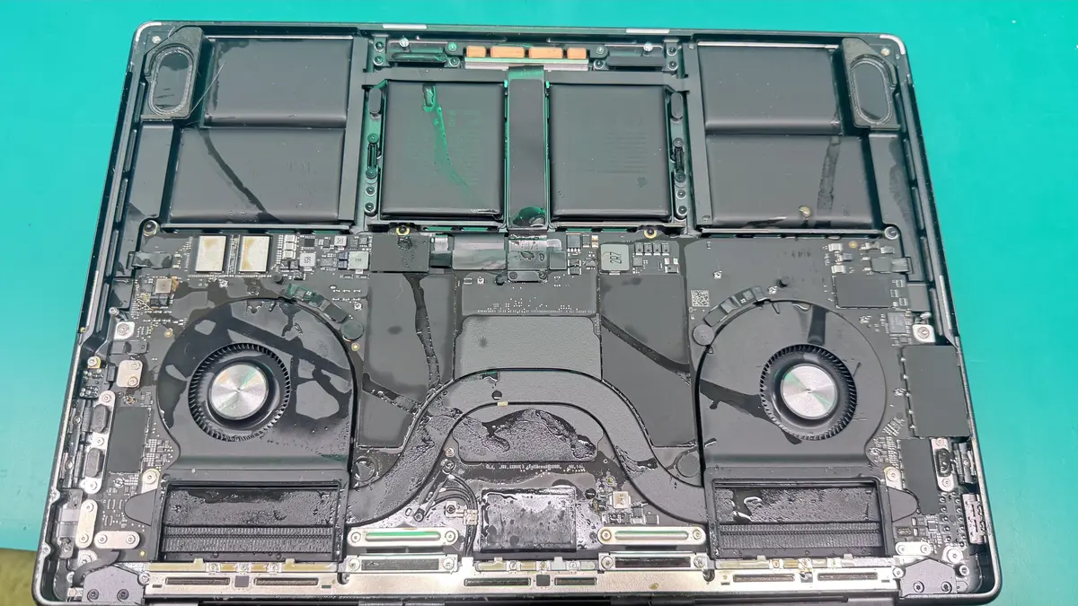 MacBook Pro with liquid damage showing corroded logic board, fans, and battery on WarriorMac workbench